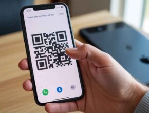 A person is holding an iPhone displaying a large black-and-white QR code on its screen, ready to be scanned. This visually demonstrates the process of using a QR code to download a mobile app. (How Do I Scan to Download an App)