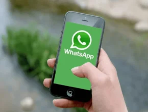 A person holding a smartphone displaying the green WhatsApp logo on the screen, with a blurred outdoor background.