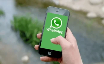 A person holding a smartphone displaying the green WhatsApp logo on the screen, with a blurred outdoor background.